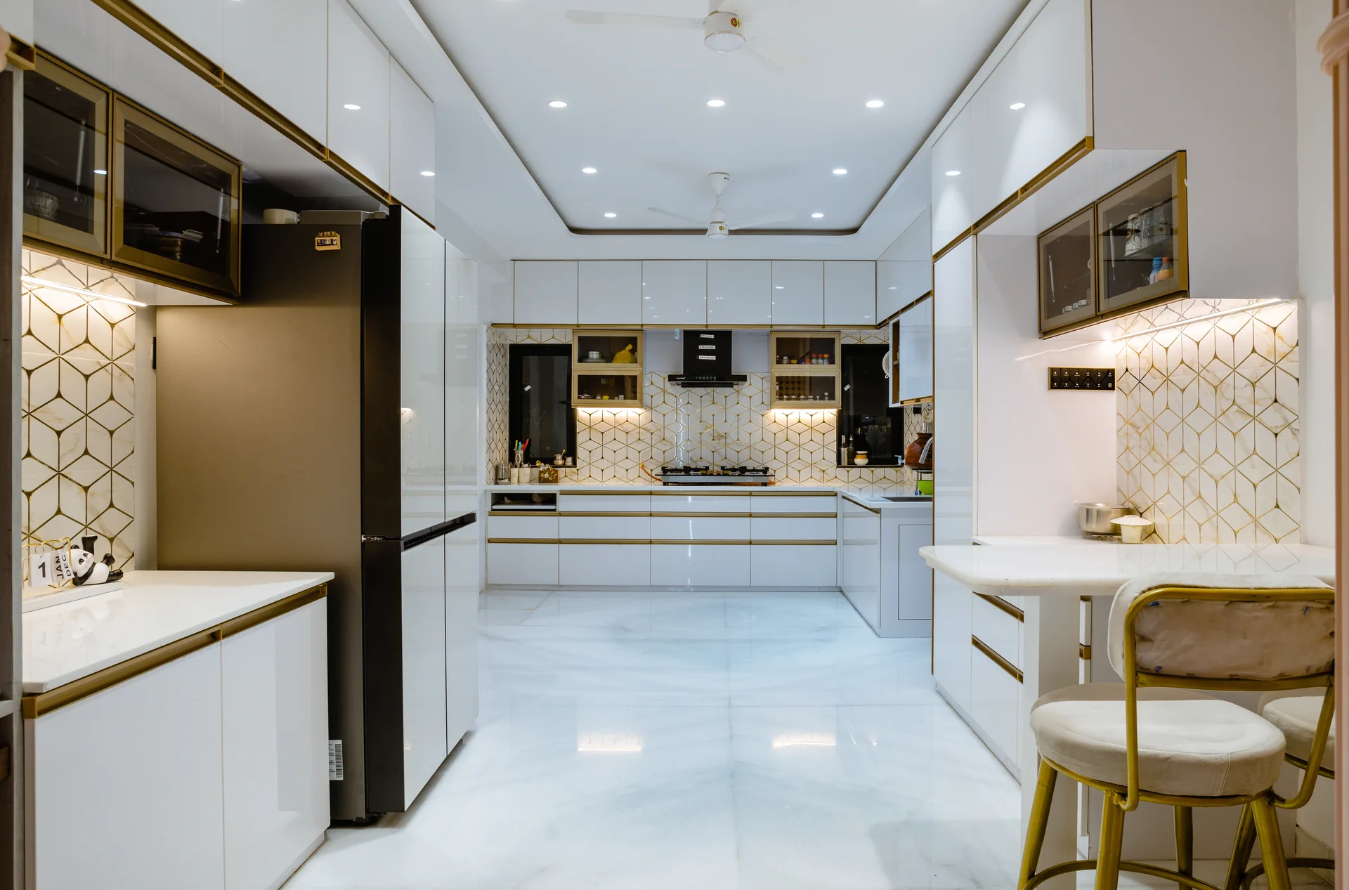 A sophisticated modular kitchen featuring crisp white cabinetry, warm metallic accents, patterned backsplash, integrated lighting, and a breakfast counter, balancing elegant aesthetics with everyday functionality.